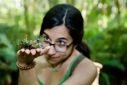 student with black hair inspecting a tiny mushroom with forest in background