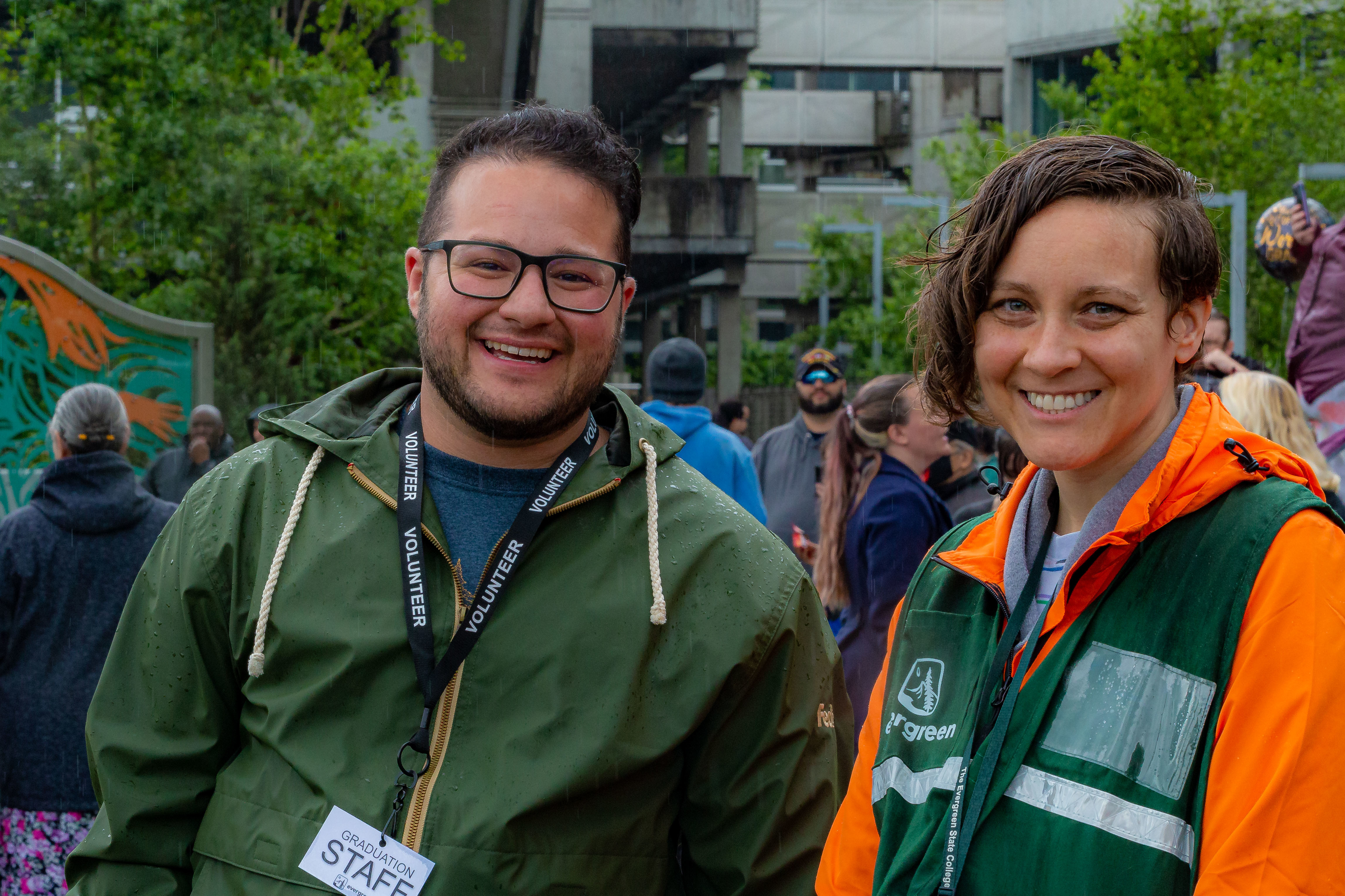 Two staff members smiling outside. One with green jacket. The other with green vest and orange jacket underneath.