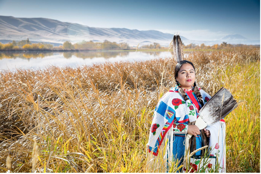 Emily Washines a tribal member in traditional dress, stands in a field near a river with mountains behind them. 