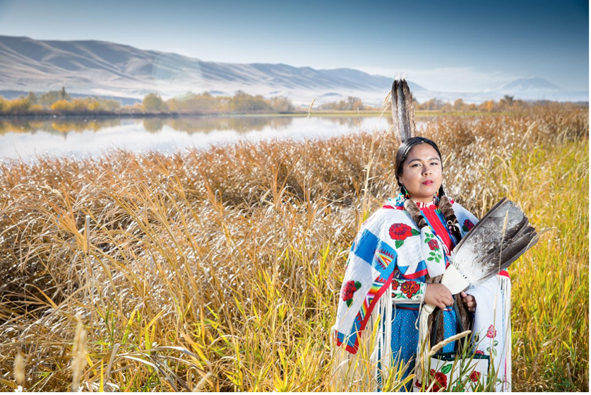 Emily Washines a tribal member in traditional dress, stands in a field near a river with mountains behind them.