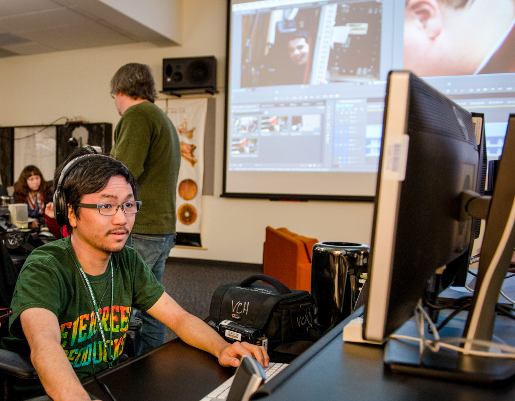 Student sits at a desk, editing a video on a computer, while the editing program is projected on a wall in the background