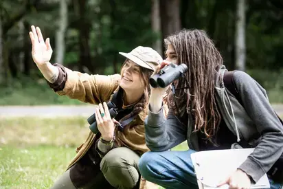 Two students crouching down in a field with binoculars, one student has their arm outstretched pointing up towards the sky