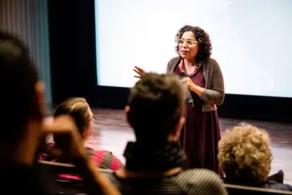 A woman standing at the front of the room with a microphone speaking, we are looking through as if we are sitting with the group.