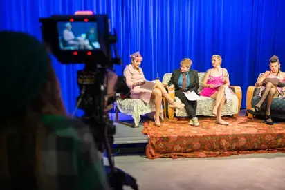 looking at four students on a stage with scripts, off to the left is a camera monitor