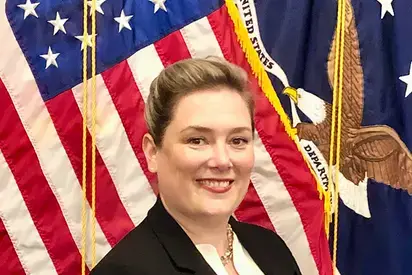 Christine M. Carlson stands for a portrait in front of the American Flag. She has fair skin, her blonde hair is pulled back, and she is wearing a white blouse with a black blazer. 