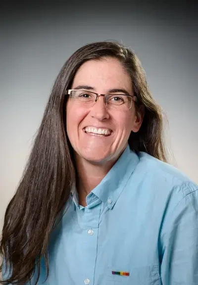 a white woman in a blue button down shirt with long brown hair and glasses, she is smiling at the camera