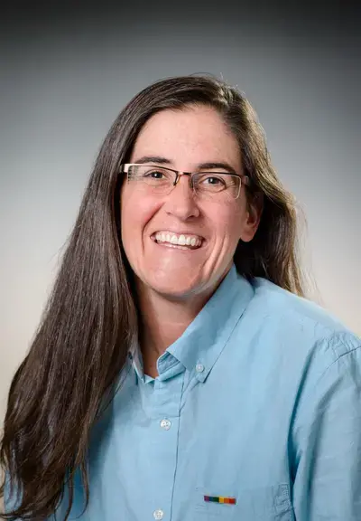 a white woman in a blue button down shirt with long brown hair and glasses, she is smiling at the camera