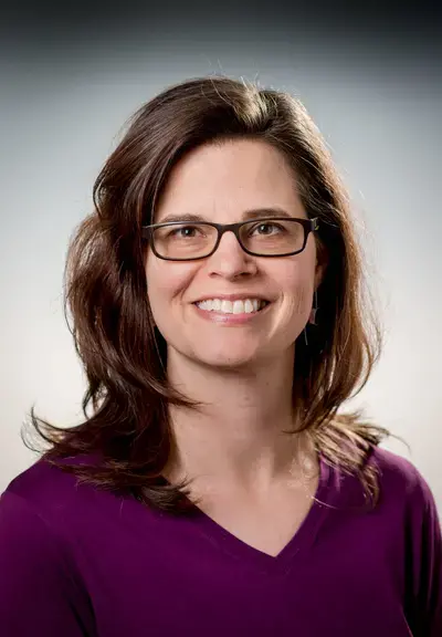 a white woman with shoulder length brown hair and glasses smiling at the camera, she is wearing a purple v-neck sweater