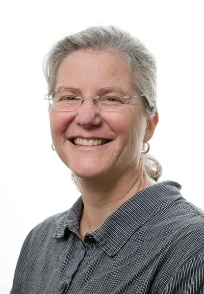 a white woman with gray hair pulled back into a ponytail, she is wearing glasses and a gray shirt and smiling at the camera