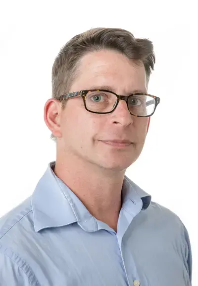 a white man with short brown hair and glasses looking at the camera, he is wearing a light blue shirt