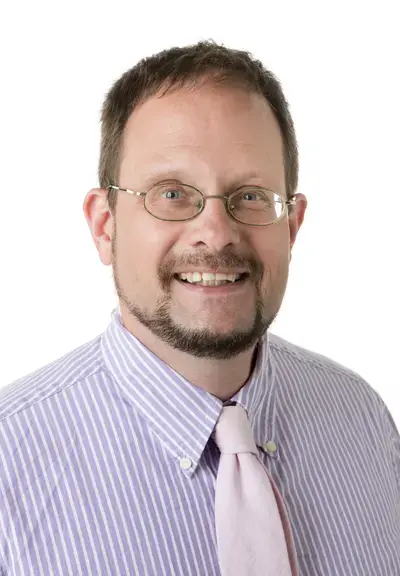 a white man with glasses, short cropped brown hair, and a trimmed beard. he is wearing a light purple pinstriped shirt and a white tie.