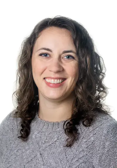 a photo of a white woman with curly brown hair and a grey sweater on, she is smiling at the camera