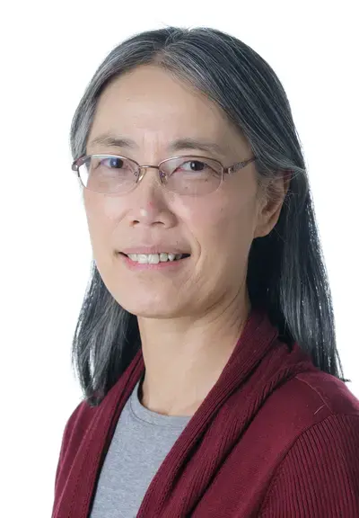 a Chinese woman with glasses and shoulder-length gray hair smiling at the camera, she is wearing a maroon cardigan with a grey shirt