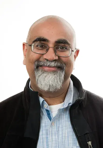 headshot of Krishna Chowdary, they have brown skin and are bald with a grey beard, they are wearing glasses and smiling at the camera