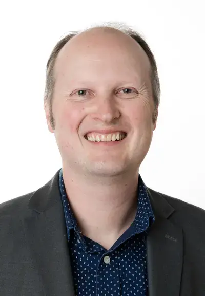 a white man smiling at the camera, he is wearing a blue polka dot button down and a black blazer