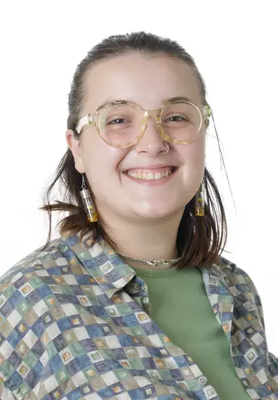 a white person with shoulder length brown hair pulled back, they are wearing glasses and dangly earrings with a green shirt and a checkered button down. they are smiling at the camera