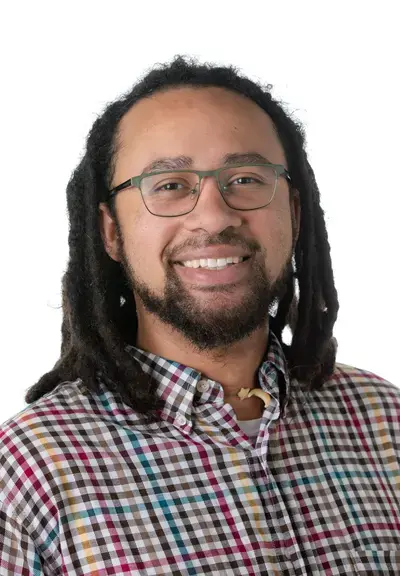 a black man with shoulder length dreads and a short beard smiling at the camera, he is wearing glasses and a multi-colored checkered shirt