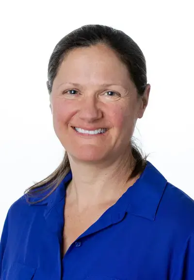 a white woman with brown hair pulled back, they are wearing a blue button down