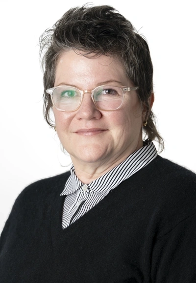 Person with short dark hair wearing glasses and a black sweater over a black and white striped buttun up shirt