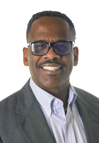 headshot of a person with short dark curly hair wearing dark rimmed glasses and a grey sports coat over a light grey button up shirt