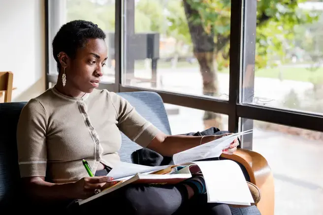 a black woman with cropped hair sits in a chair by a window going through a notebook