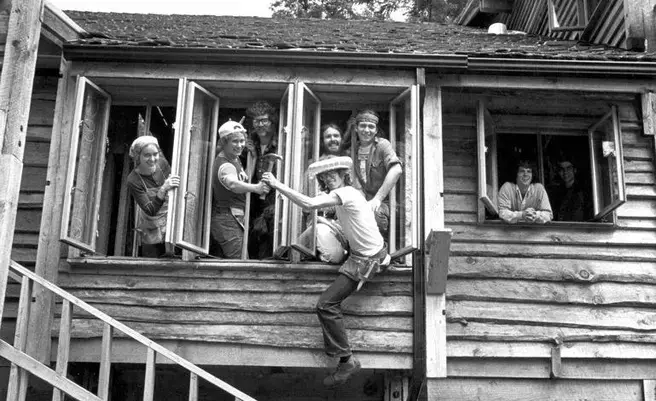 a black and white photo of students hanging out the windows of the farm house, one student is hanging off over the stairs out of the window sill, holding on to peoples hands inside the building