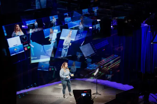 student on stage with a small melon, multiple screens and projections behind them on stage