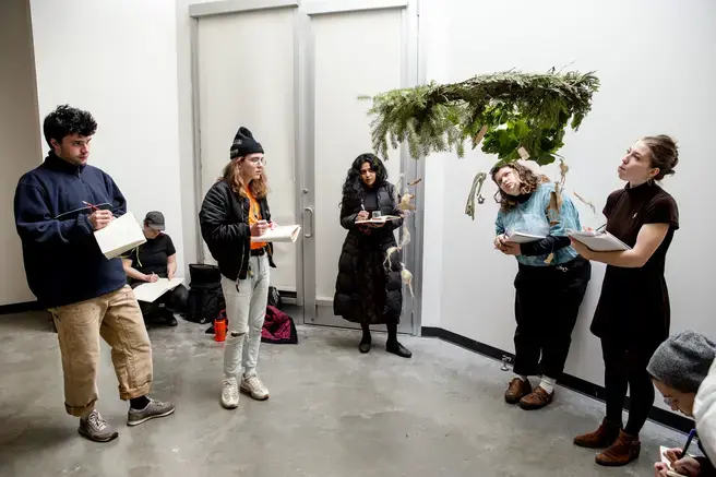 A group of students stands around a found-object sculpture suspended from the ceiling.
