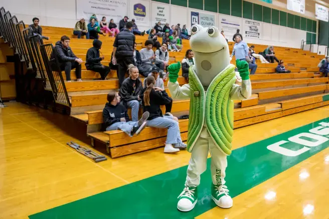 Speedy the geoduck standing in front of people sitting in the bleachers in a gym