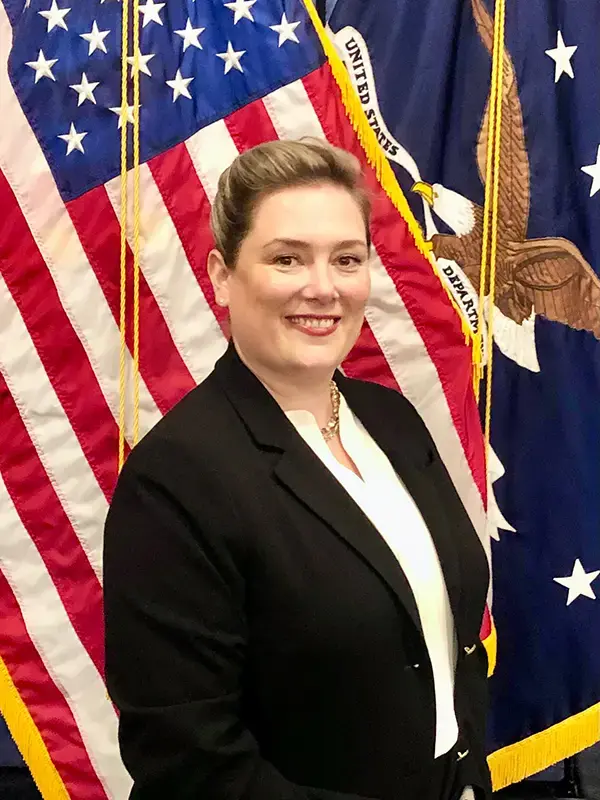 Christine M. Carlson stands for a portrait in front of the American Flag. She has fair skin, her blonde hair is pulled back, and she is wearing a white blouse with a black blazer. 