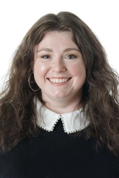 a white woman with long brown wavy hair smiling a the camera, she is wearing a dark sweater with a white collar coming out the top