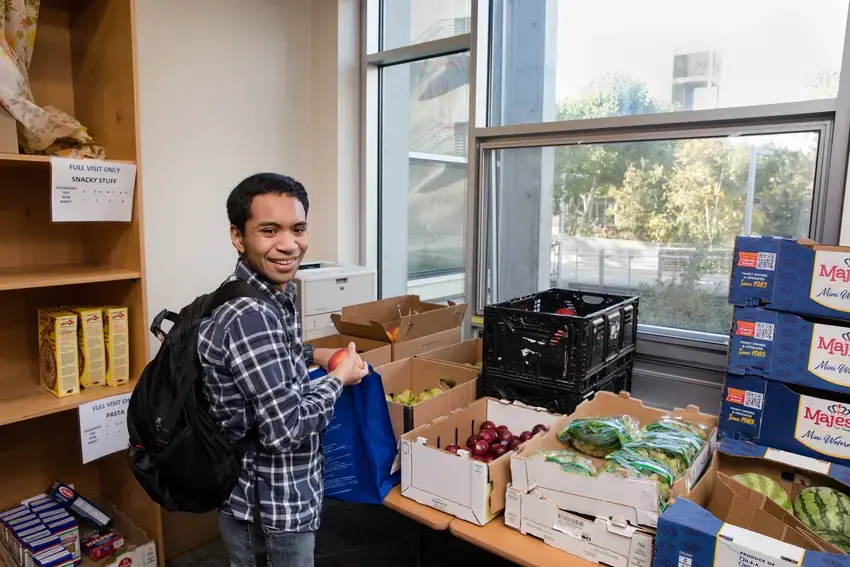 A student filling a back at the foodbank, looking back over their shoulder at the camera and smiling. they are holding an apple. 