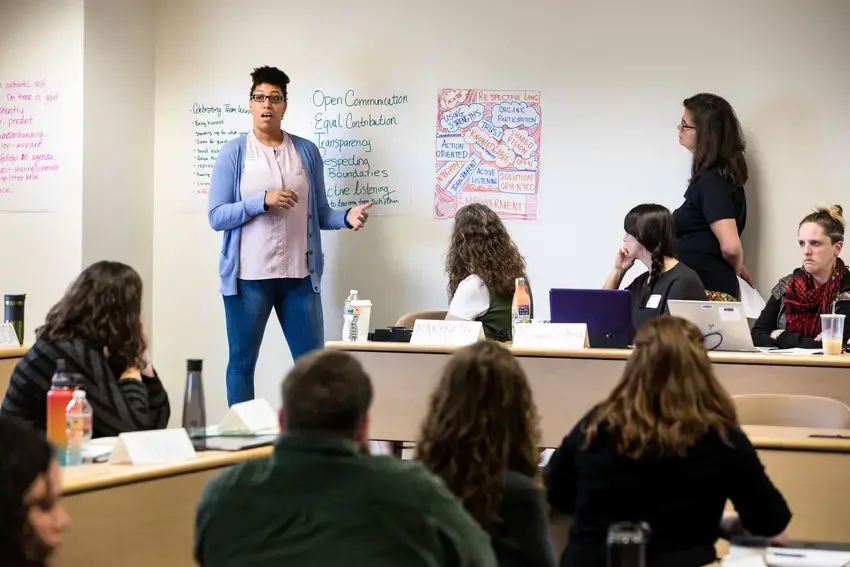 an mpa student leading a class discussion. they are gesturing to the wall behind them and making notes on a large piece of paper