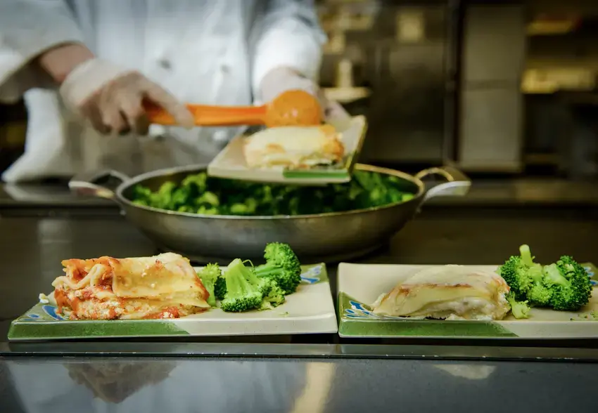 Close up of dishes of pasta and broccoli with out of focus hands in the background filling another dish