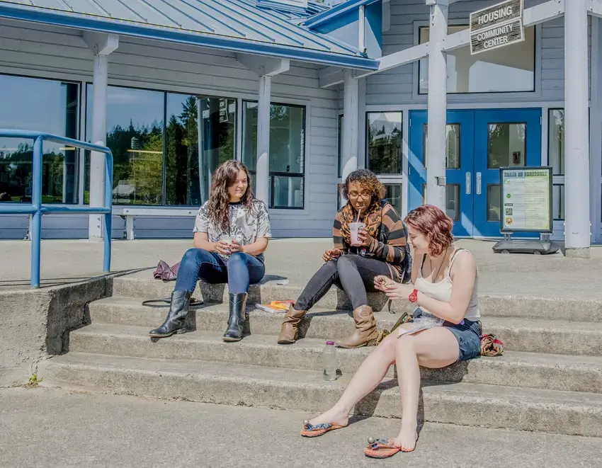 Three students sit on the stepa of the housing community center enjoying the sun