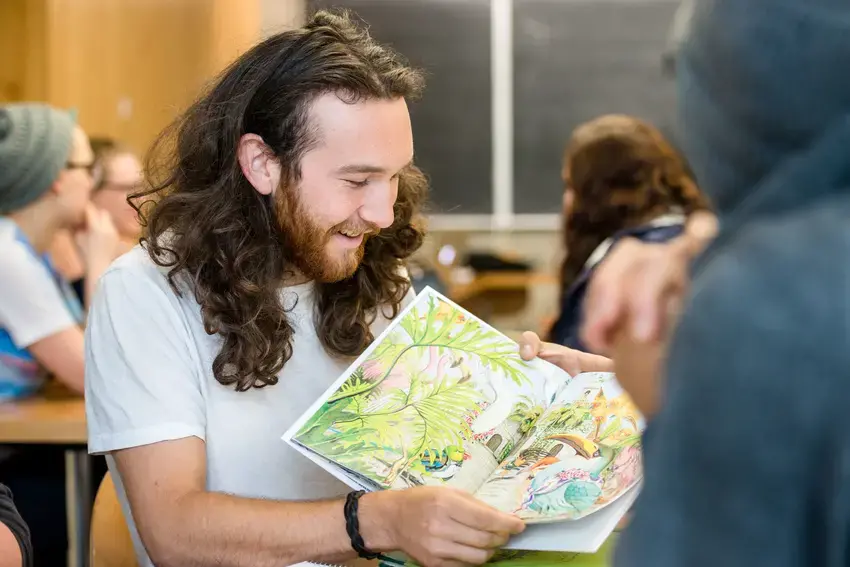 a young man with long hair holding a children's book out as if reading to a group