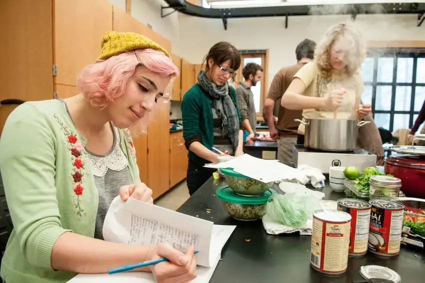 a student is sitting near the camera going over their notes, other students are in the background cooking over a large pot. cans of food and produce are on the counter. 