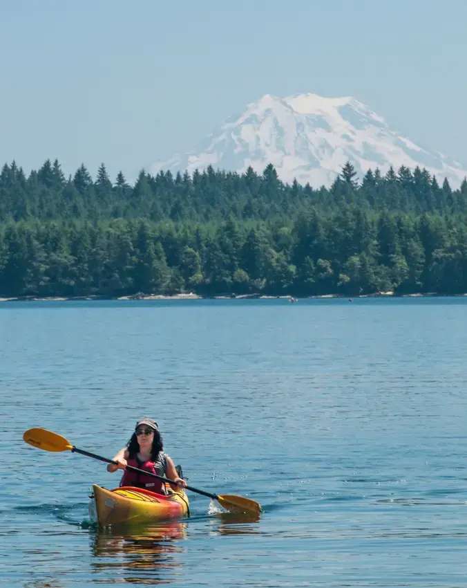 A person in a yellow kayack on the water with trees and a view of Mt Rainier in the background