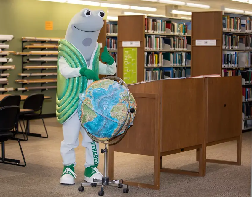 speedy the geoduck mascot standing behind a large globe in the library and giving a thumbs up
