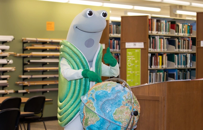 speedy the geoduck mascot standing behind a large globe in the library and giving a thumbs up