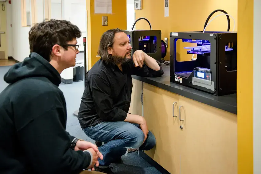 two people squatting to look inside of a 3D printer in the CAL 