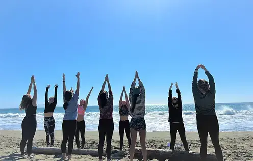 Sierra Wagner teaching yoga to a group of women on the beach, the sky is clear blue, and waves are crashing behind them.