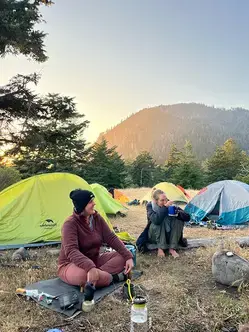 Two retreat participants sit around a campfire chatting with others (out of frame) in the mountains at sunrise, there are 6 tents set up in the background.
