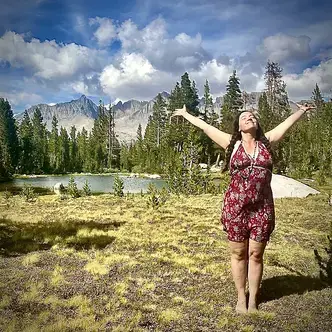 Natalie Pyrooz stands near an alpine lake in the mountains with her arms stretched wide and head looking up to the sky.​