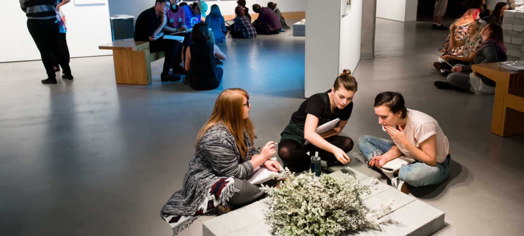 Three female students with notebooks sit close to a piece of artwork made of wood and flowers on the floor, holding a discussion, inside an art gallery. Other students are visible in the background doing the same.