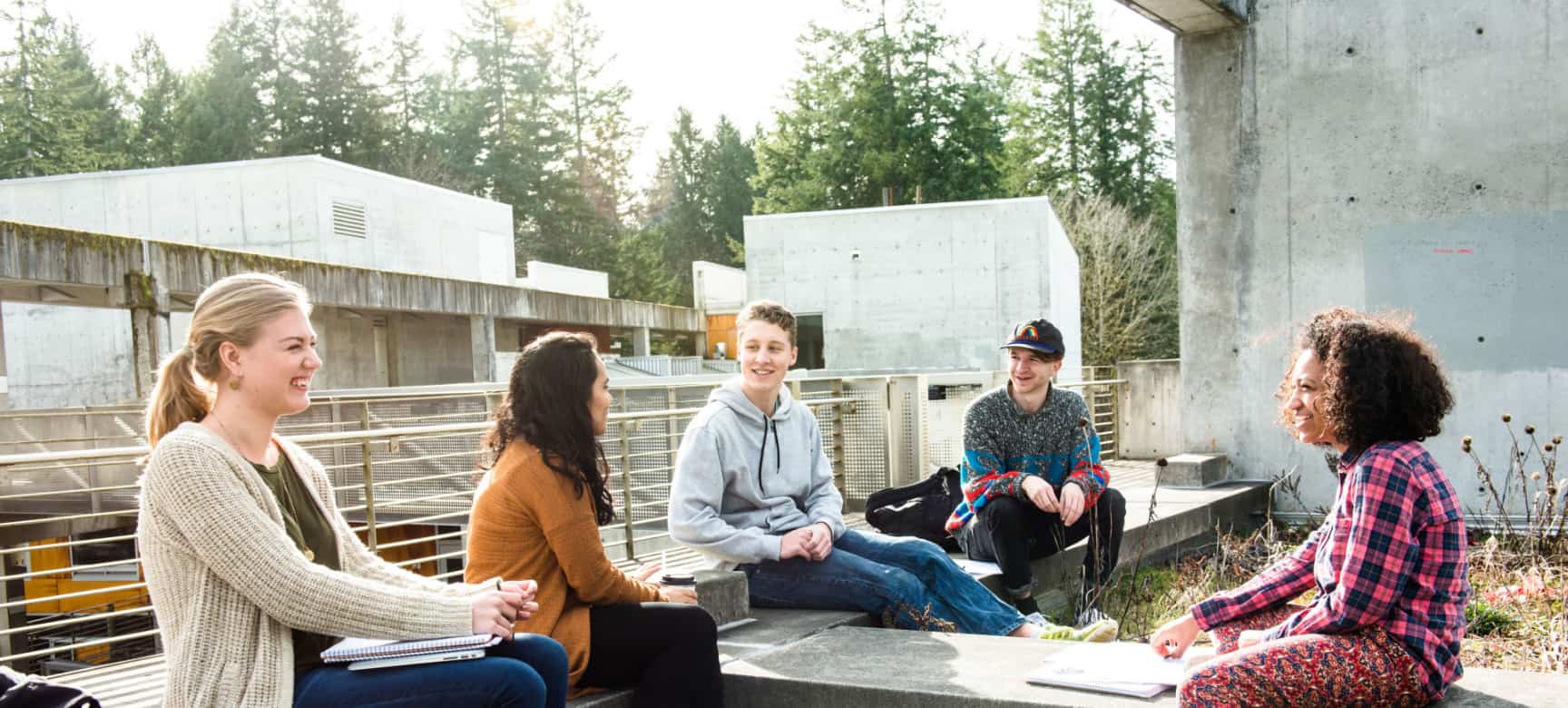 Five students of different races and backgrounds, 2 male and 3 female, sit outside on an overgrown loading dock laughing and discussing topics. Trees and industrial buildings are visible in the background.