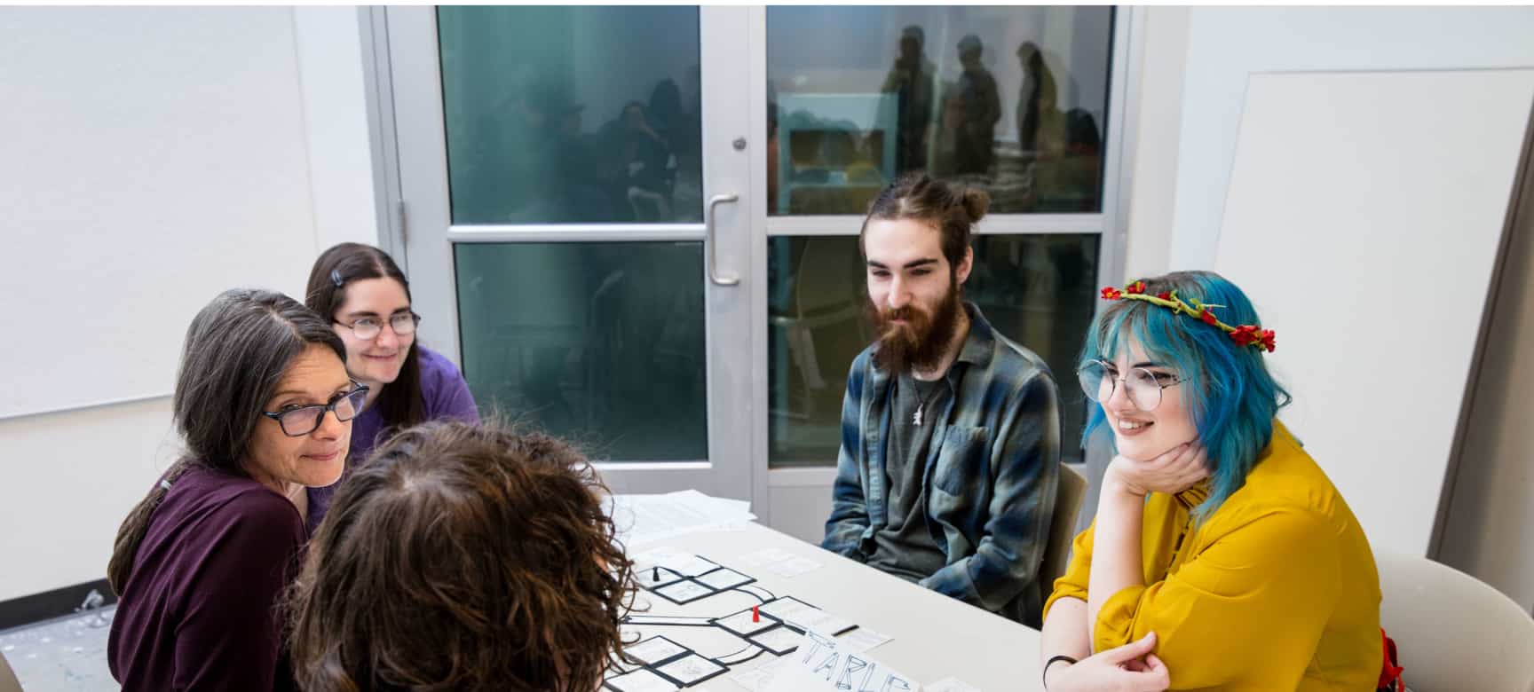 Four students, 3 female and one male, sit around a white table with a female teacher discussing class topics