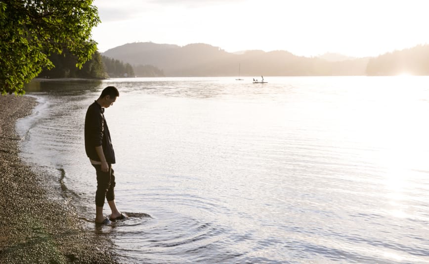 A man stands at the shore of lake with his feet in the water, brushing against the shoreline.