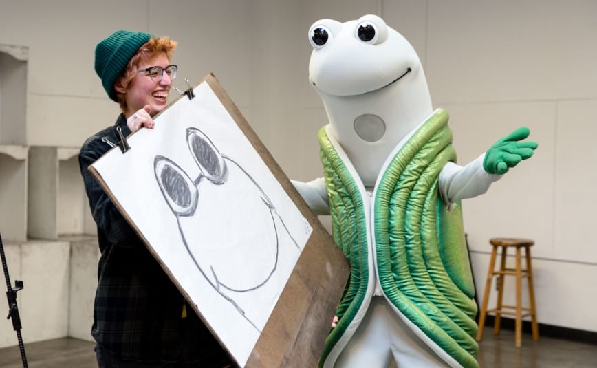 Speedy, the mascot for Evergreen, stands next to a laughing woman holding a sketch of Speedy in an art studio.