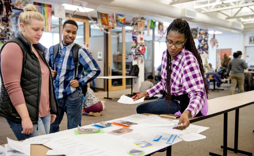 An African-American woman shows two other students, one a White female and the other an African-American male, some papers on a workbench within a classroom. The man is smiling.